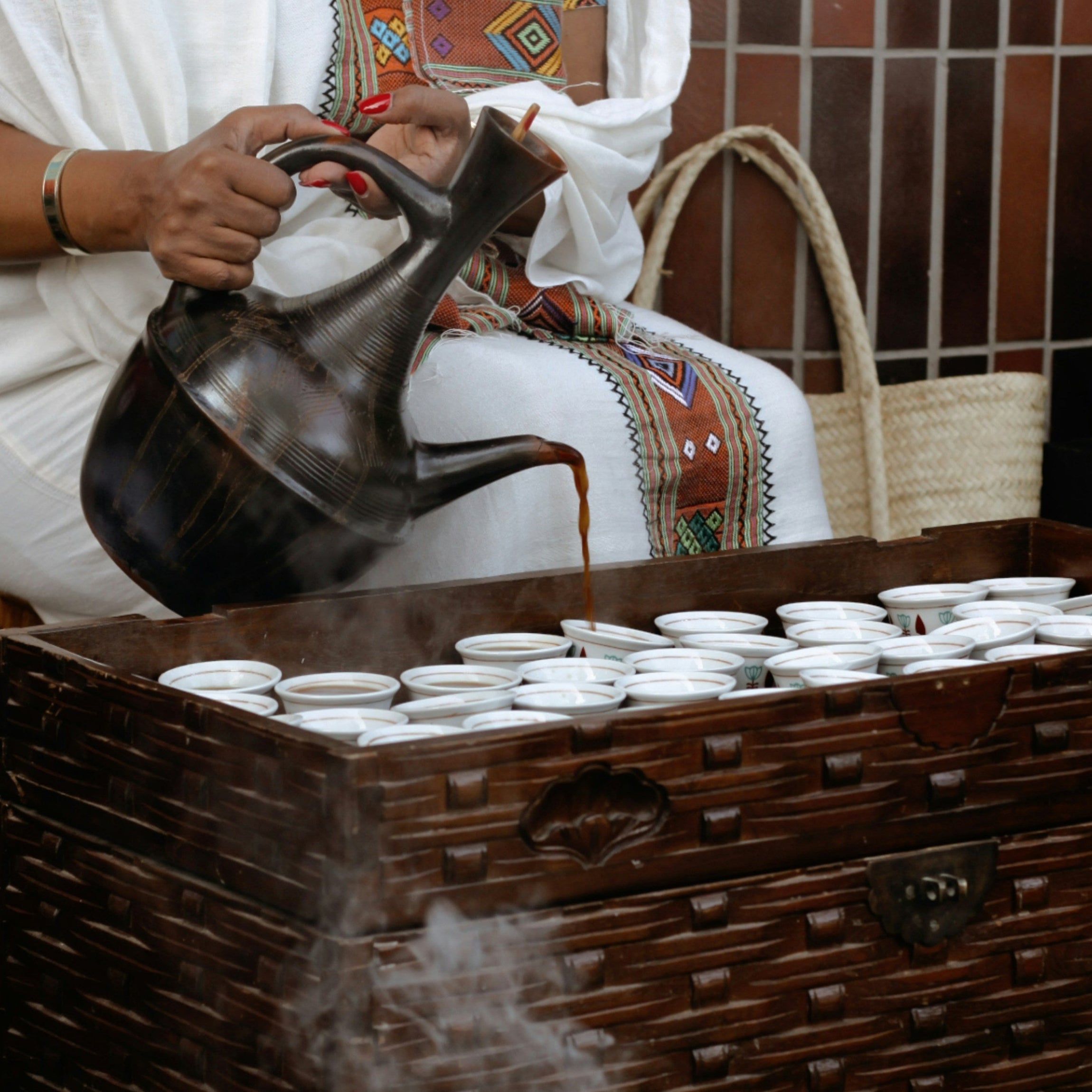 Person pouring water from a pitcher into a large wooden container with smoke rising, surrounded by baskets and a woven chair.